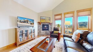A living room with a stone fireplace, a television, a bookshelf, a leather sofa, and large windows with a view. - Modern Mountain Retreat/Incredible Mountain Views/Stylish Kitchen/Spotless (Estes Park)