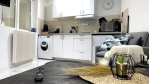A modern kitchen with white cabinets, a microwave, and a coffee maker. There is a washing machine to the left and a gray sofa with a geometric pillow in the living area.