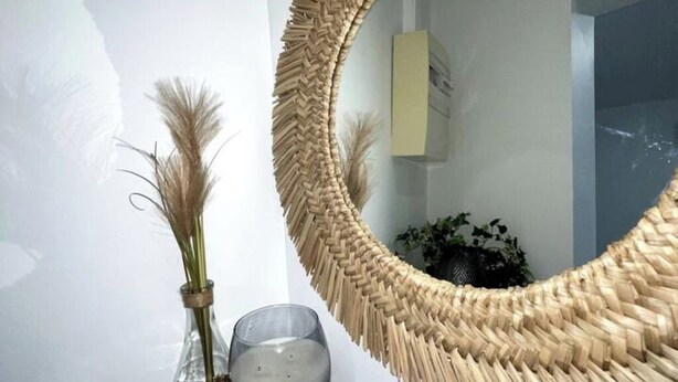 A mirror with a woven frame, a vase with pampas grass, and a candle on a surface.