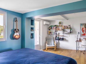 A bedroom with a bed, a guitar, a chair, a desk with a lamp, and shelves with books and decorative items.