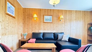 Una habitación acogedora con un revestimiento de madera, un sofá de cuero negro, dos sillas rosas, una mesa de madera y dos lámparas fijadas a la pared.