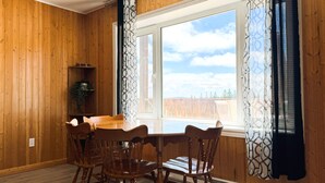 Una habitación con paneles de madera, una mesa de comedor y sillas, una ventana con cortinas estampadas y una pequeña planta en un estante.