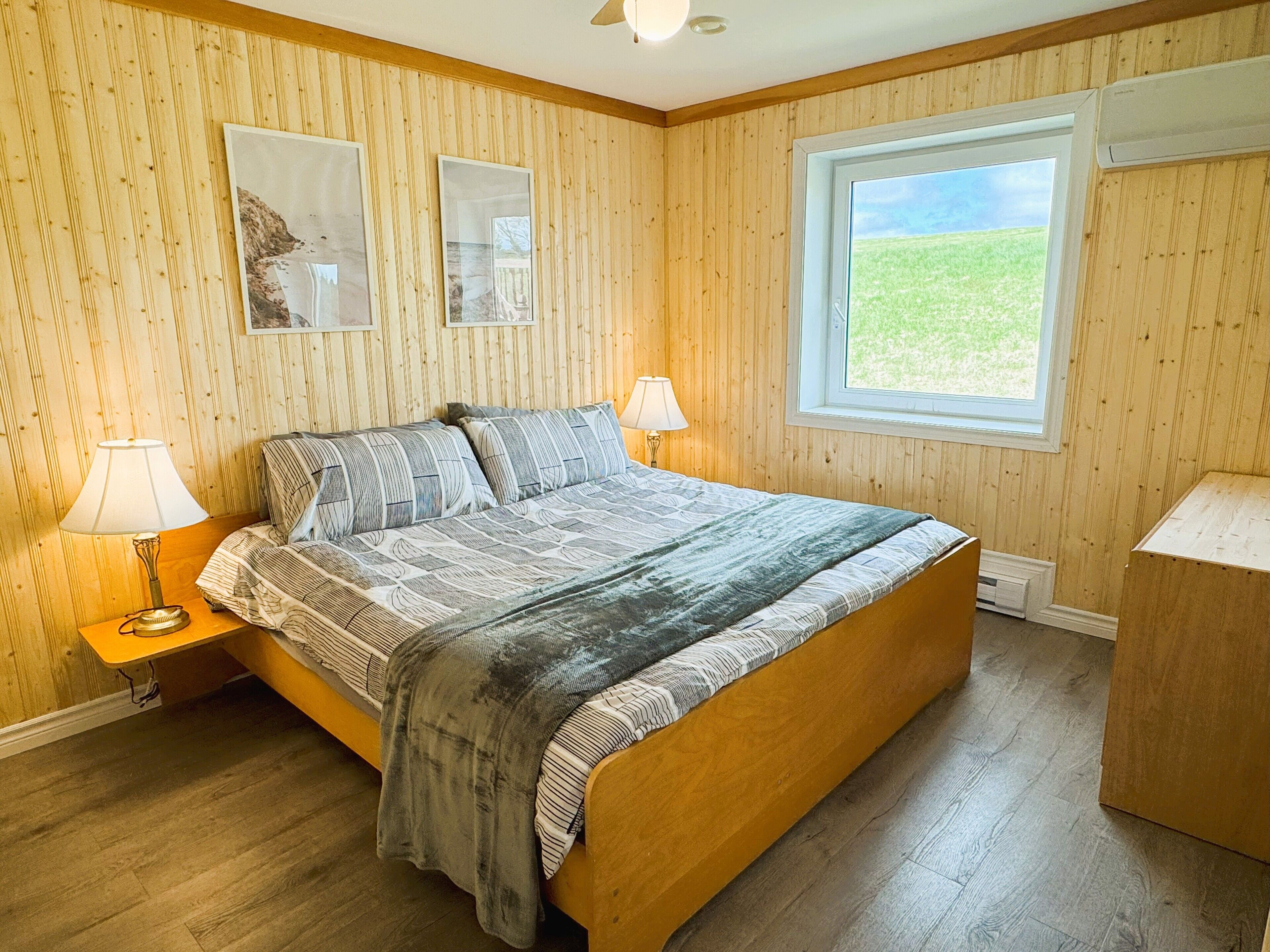A bedroom with a wooden bed, two bedside lamps, a window with a view of a grassy field, and two framed pictures on the wall.