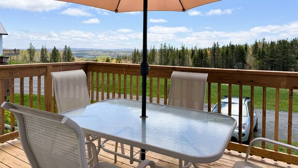 Una terraza de madera con una mesa y sillas de vidrio, una sombrilla y vistas a un bosque y montañas lejanas.