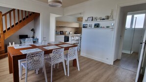 A dining area with a wooden table and white chairs, a staircase, and a kitchen in the background. - Pretty and bright renovated house in the center of Trégunc with enclosed garden (Trégunc)