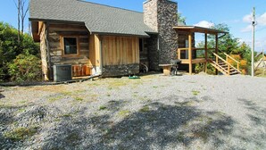 A rustic log cabin with a stone chimney, a wooden porch, and a gravel driveway.