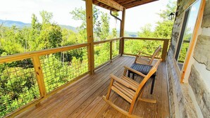 A wooden deck with a rocking chair and a table, overlooking a lush green landscape.