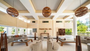 A spacious lobby with a high ceiling, large windows, and a central seating area with sofas and tables. - Luxury 2BR Condo with Pool - Paseo del Sol Playacar (Playa del Carmen)