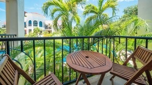 A balcony with a round wooden table and two wooden chairs, overlooking a pool and palm trees. - Luxury 2BR Condo with Pool - Paseo del Sol Playacar (Playa del Carmen)