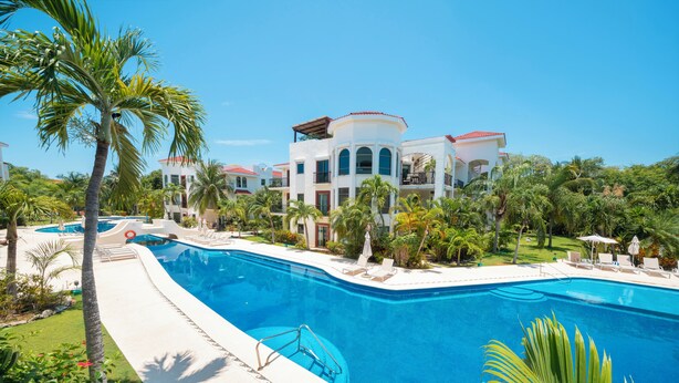 A large swimming pool surrounded by palm trees and a resort building with multiple balconies. - Luxury 2BR Condo with Pool - Paseo del Sol Playacar (Playa del Carmen)