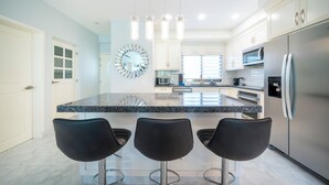 A modern kitchen with a large island, bar stools, stainless steel appliances, and a decorative clock. - Luxury 2BR Condo with Pool - Paseo del Sol Playacar (Playa del Carmen)