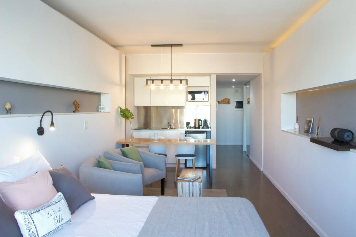 A modern hotel room with a bed, a seating area, a kitchenette, and a small dining table.