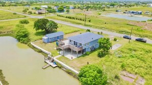 Una casa de dos pisos con un muelle que se adentra en un cuerpo de agua, rodeada de campos abiertos y algunos árboles dispersos.