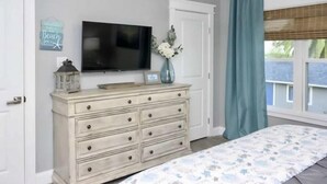 Un dormitorio con un aparador, televisor, un florero con flores y una ventana con cortinas.