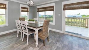 Un comedor con una mesa y sillas, ventanas grandes con persianas y vistas a una terraza y follaje exterior.