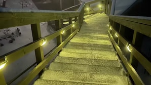 A staircase with yellow railings leading to a building with a snow-covered roof and a lit window.