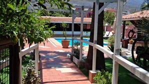 En sti der fører til et poolområde med en pergola, omgivet af plantekasser og udsigt til bygninger i baggrunden.