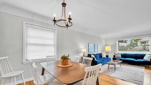 A modern living room with a dining area, featuring a wooden dining table, white chairs, a blue sofa, and a chandelier. - Elegant, Artsy Home in N. Royal Oak! (Royal Oak)