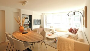 A modern living room with a round wooden dining table, beige sofas, and a large window providing natural light. - Apartment in Het Zoute Near the Beach (Knokke-Heist)