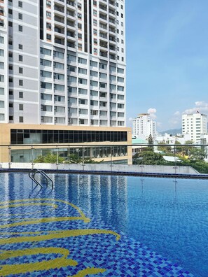 A modern hotel with a large pool area, featuring a clear blue pool with yellow and blue mosaic patterns, surrounded by a glass facade building.