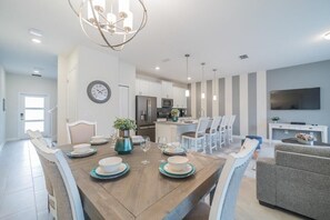 Una cucina moderna con elettrodomestici in acciaio inossidabile, una zona pranzo con tavolo in legno apparecchiata per quattro persone e un soggiorno con divano grigio e televisore a schermo piatto.
