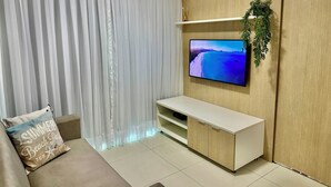 A modern living room with a flat-screen TV mounted on a wooden wall, a beige sofa with a decorative pillow, and a large window with sheer curtains.