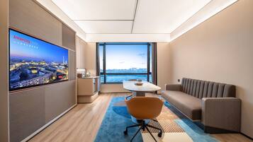 Ein modernes Hotelzimmer mit großem Fernseher, der eine Stadtansicht zeigt, einer bequemen Couch, einem kleinen Tisch und Blick auf die Stadt durch große Fenster.