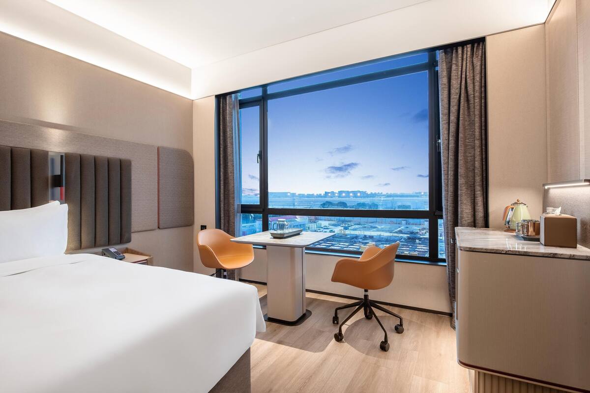 Ein Hotelzimmer mit großem Fenster, einem Bett, einem Schreibtisch, zwei Stühlen und einer Minibar.