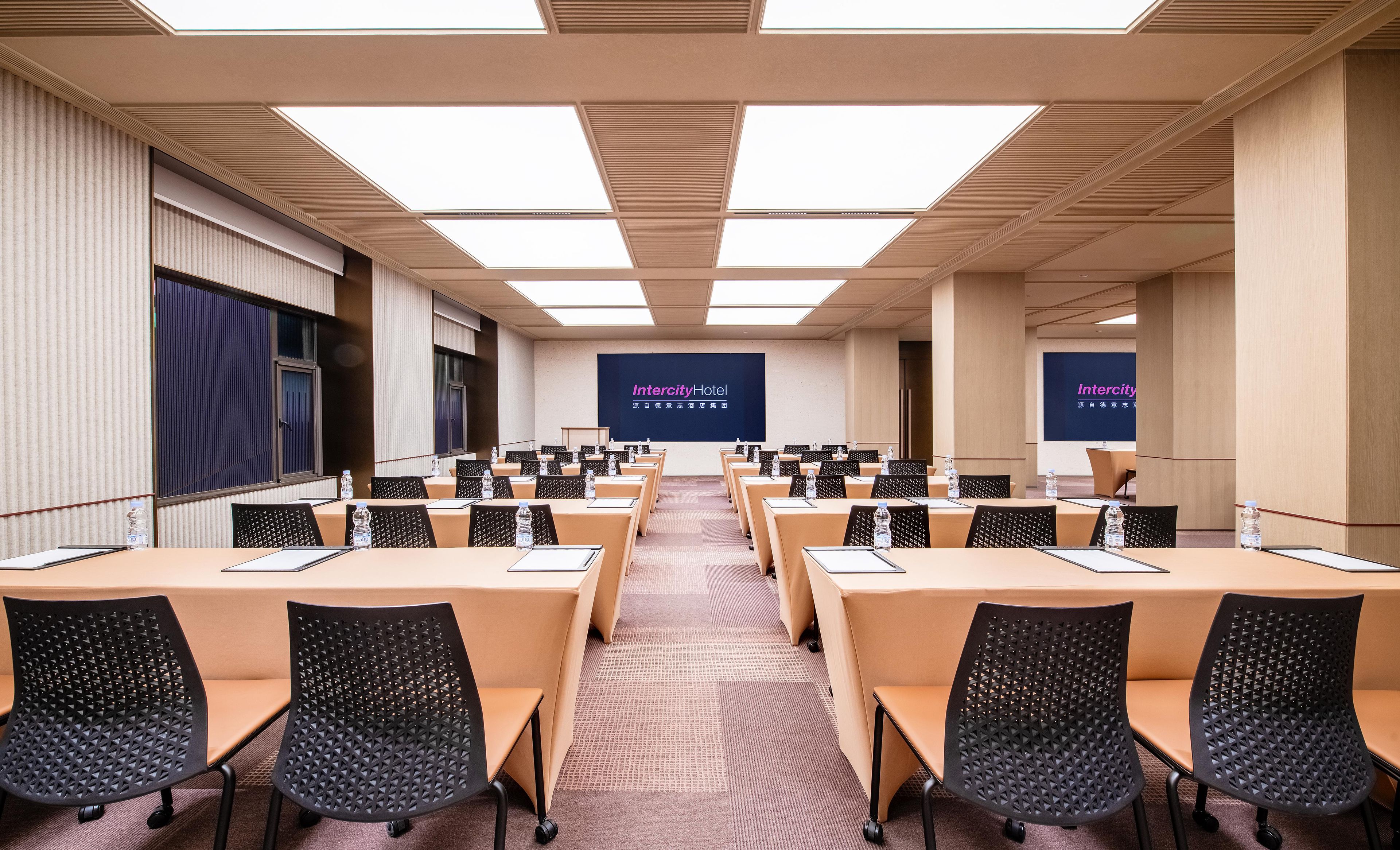 Un salón de conferencias con varias mesas y sillas, una pantalla grande mostrando la marca 'Intercity Hotel' y un diseño de techo moderno.