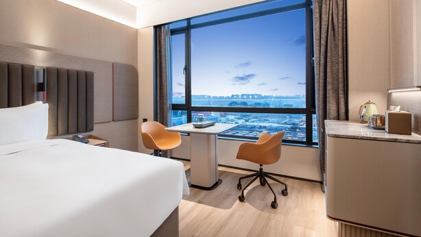 A hotel room with a large window, a bed, a desk, two chairs, and a minibar. - Intercity Hotel Shanghai Hongqiao Airport (Shanghai)