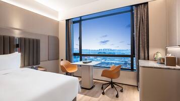 A hotel room with a large window, a bed, a desk, two chairs, and a minibar.