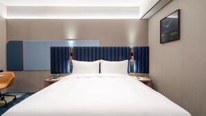 A modern hotel room with a large bed, a desk with a chair, a telephone, and a wall-mounted TV.