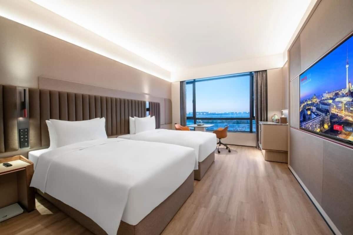 Ein Hotelzimmer mit zwei Betten, einem großen Fenster mit Blick auf die Stadt, einem Flachbildfernseher und einem kleinen Tisch.