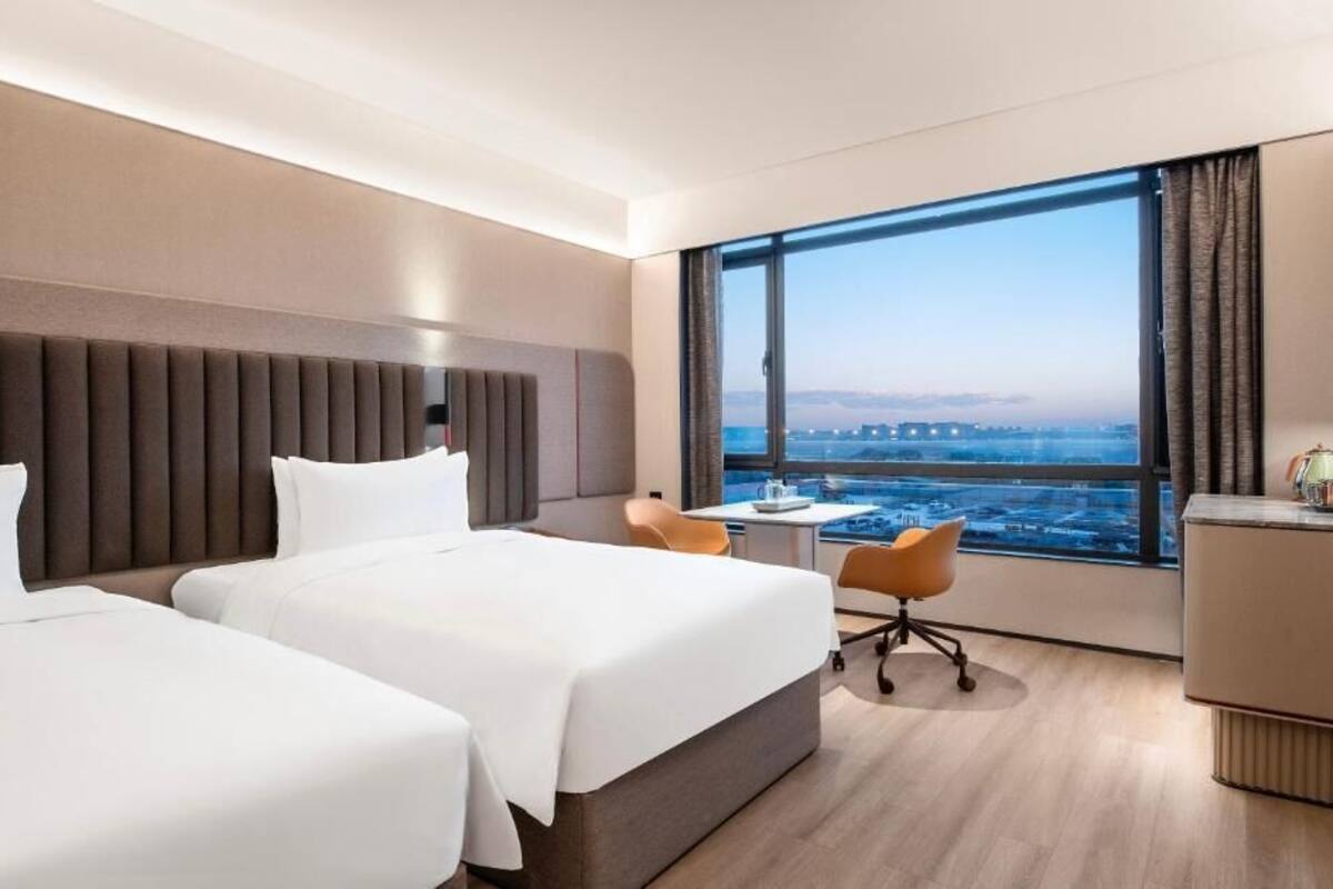 Ein Hotelzimmer mit zwei Betten, einem Schreibtisch, einem Stuhl und einem großen Fenster mit Blick auf die Stadt.