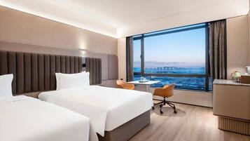 A hotel room with two beds, a desk, a chair, and a large window offering a city view.