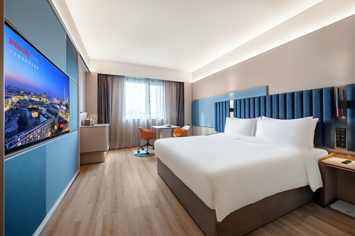 Ein modernes Hotelzimmer mit einem großen Bett, einem Schreibtisch, zwei Sesseln, einem Fernseher und Blick auf die Stadt.