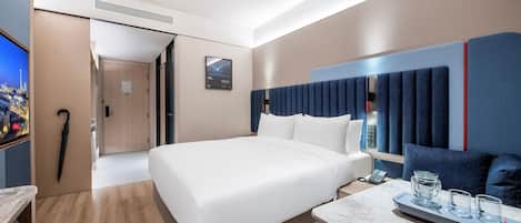 A modern hotel room with a large bed, a desk with a phone and glasses, and a TV on the left.
