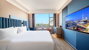 A hotel room with a large bed, a desk with a chair, a TV displaying a cityscape, and a view of the city through the window. - Intercity Hotel Shanghai Hongqiao Airport (Shanghai)