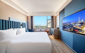 Habitación de hotel con una cama grande, un escritorio con silla, un televisor mostrando un paisaje urbano y vistas de la ciudad a través de la ventana.