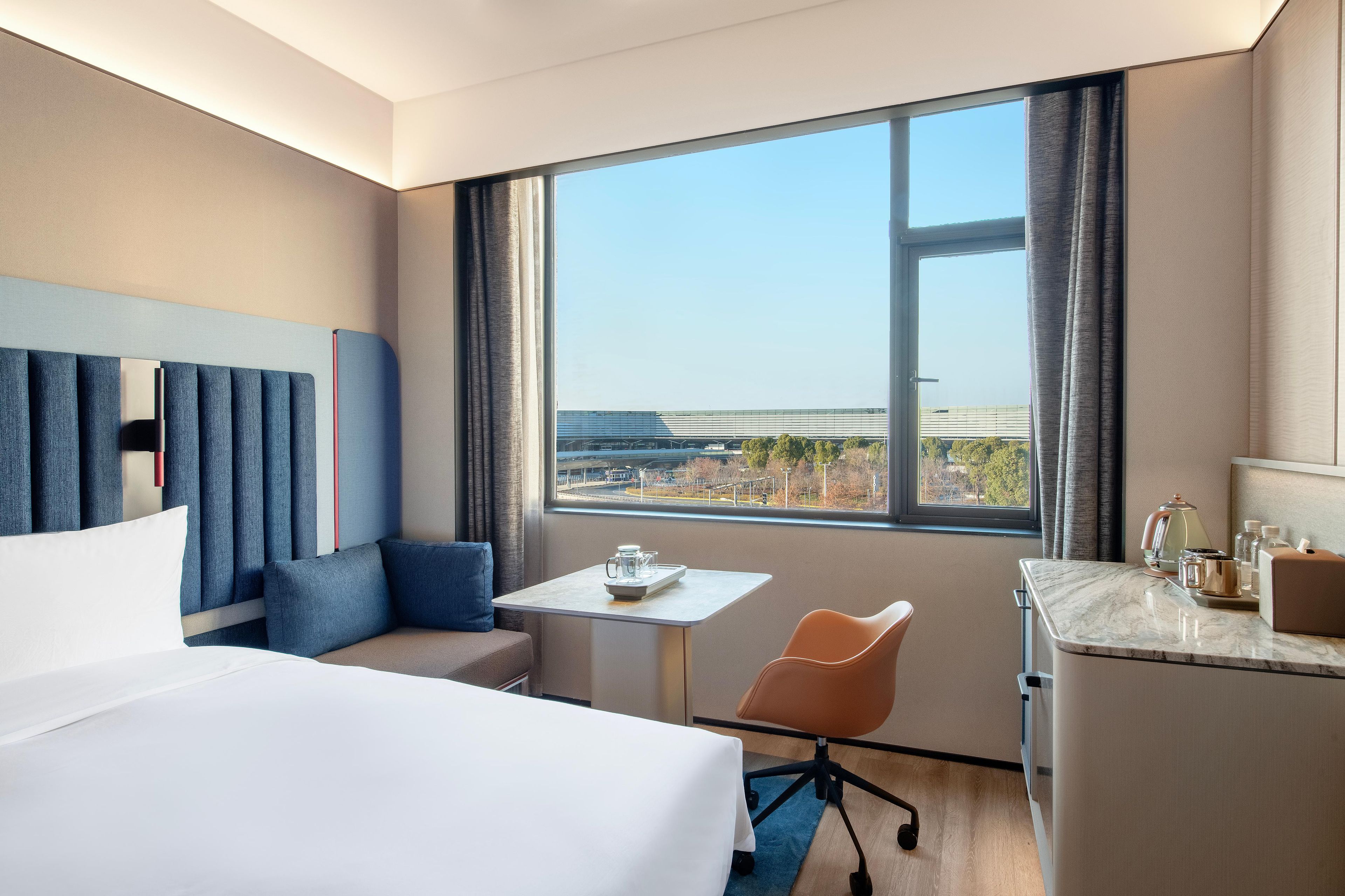 A hotel room with a large bed, a small table, a desk, a chair, and a view of the city through a large window.