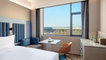 Quarto de hotel com uma cama grande, uma mesinha, escrivaninha, cadeira e vista da cidade por uma janela ampla.