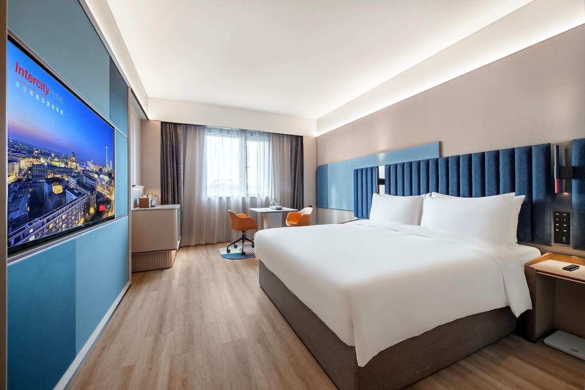 Ein modernes Hotelzimmer mit einem großen Bett, einem Schreibtisch, zwei Sesseln, einem Fernseher und Blick auf die Stadt.