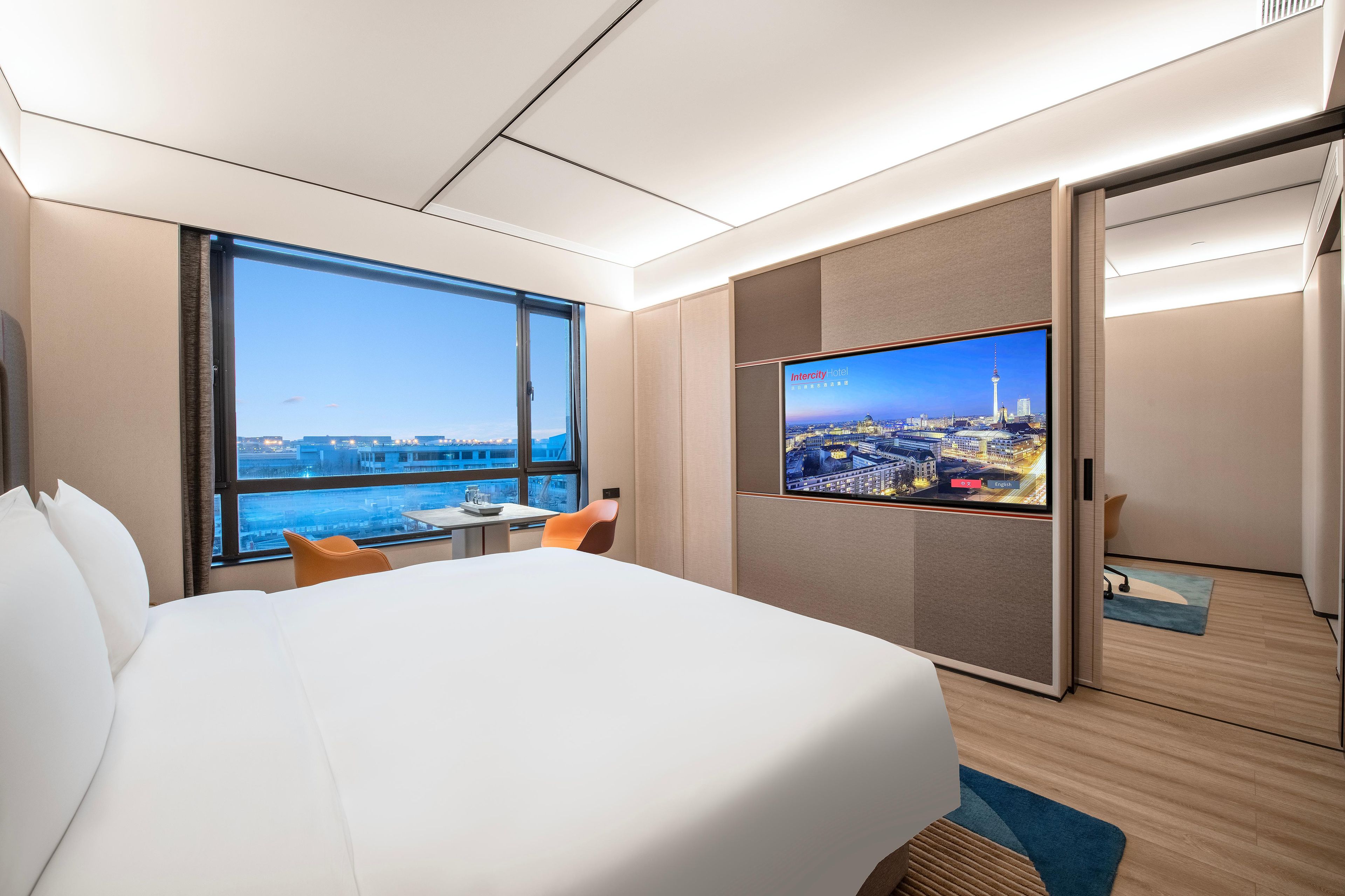 Quarto de hotel com uma cama, uma televisão exibindo vista urbana, uma janela com vista para edifícios e uma porta.