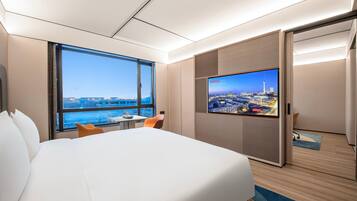 Quarto de hotel com uma cama, uma televisão exibindo vista urbana, uma janela com vista para edifícios e uma porta.