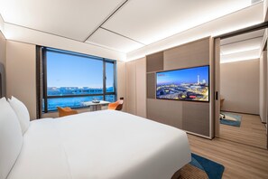 Quarto de hotel com uma cama, uma televisão exibindo vista urbana, uma janela com vista para edifícios e uma porta.