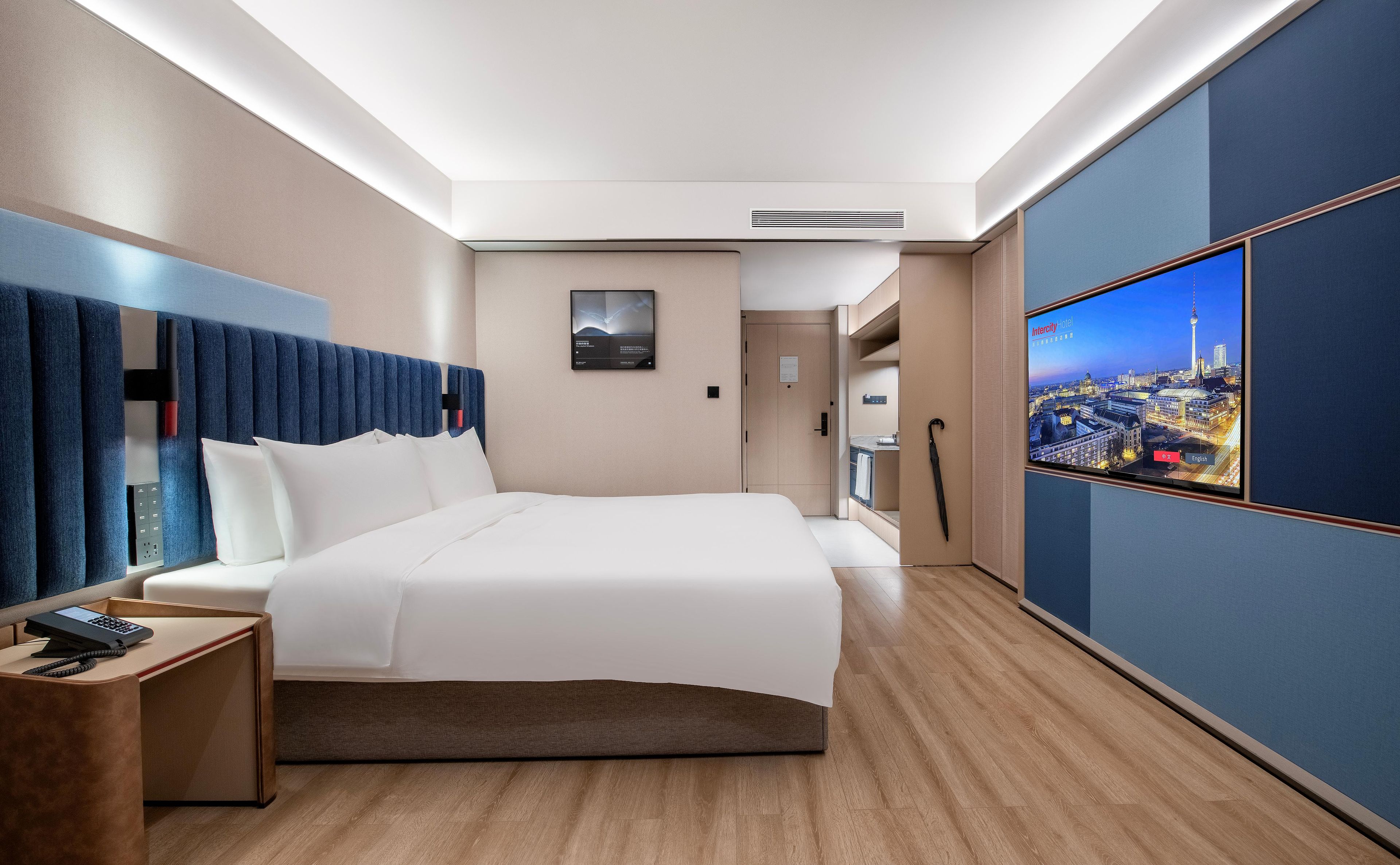 Habitación de hotel con una cama grande, un escritorio con teléfono, un televisor mostrando una vista urbana y una puerta que da al baño.