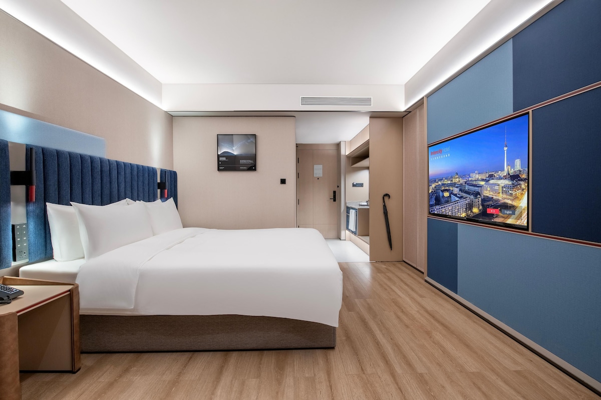 A hotel room with a large bed, a desk with a phone, a TV displaying a cityscape, and a door leading to a bathroom.