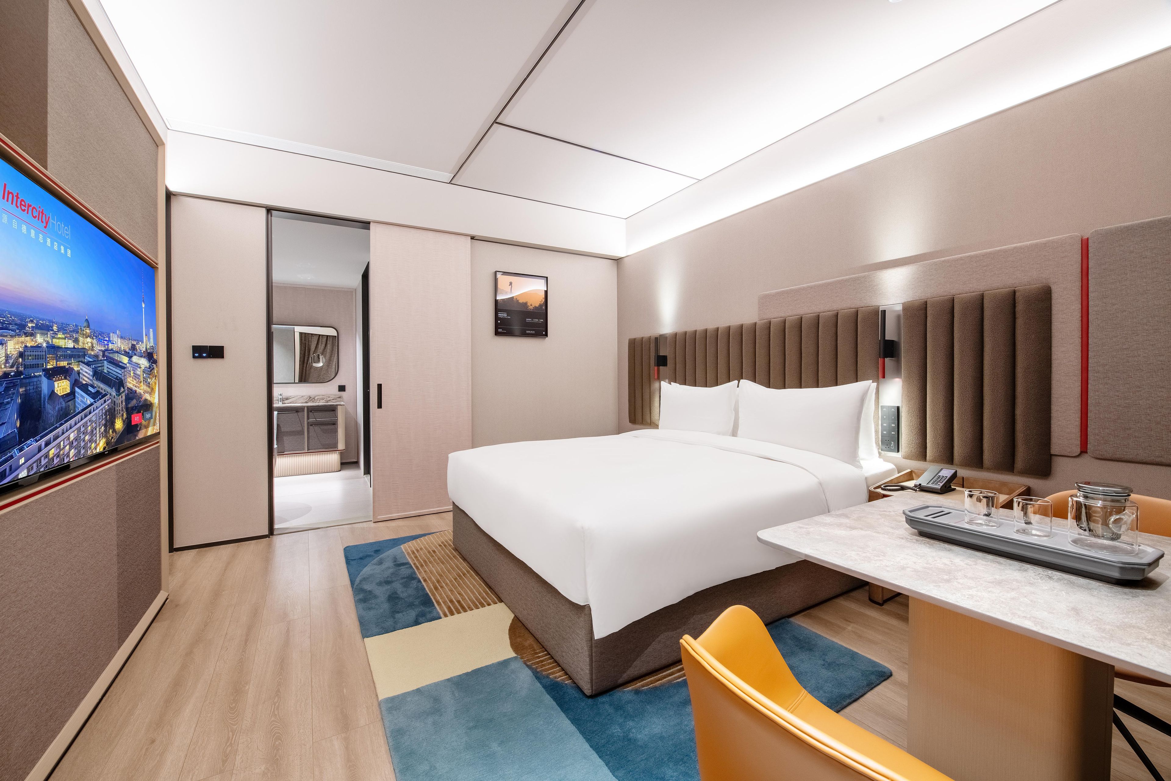 Una habitación de hotel moderna con una cama grande, un escritorio con teléfono y un televisor que muestra un paisaje urbano.