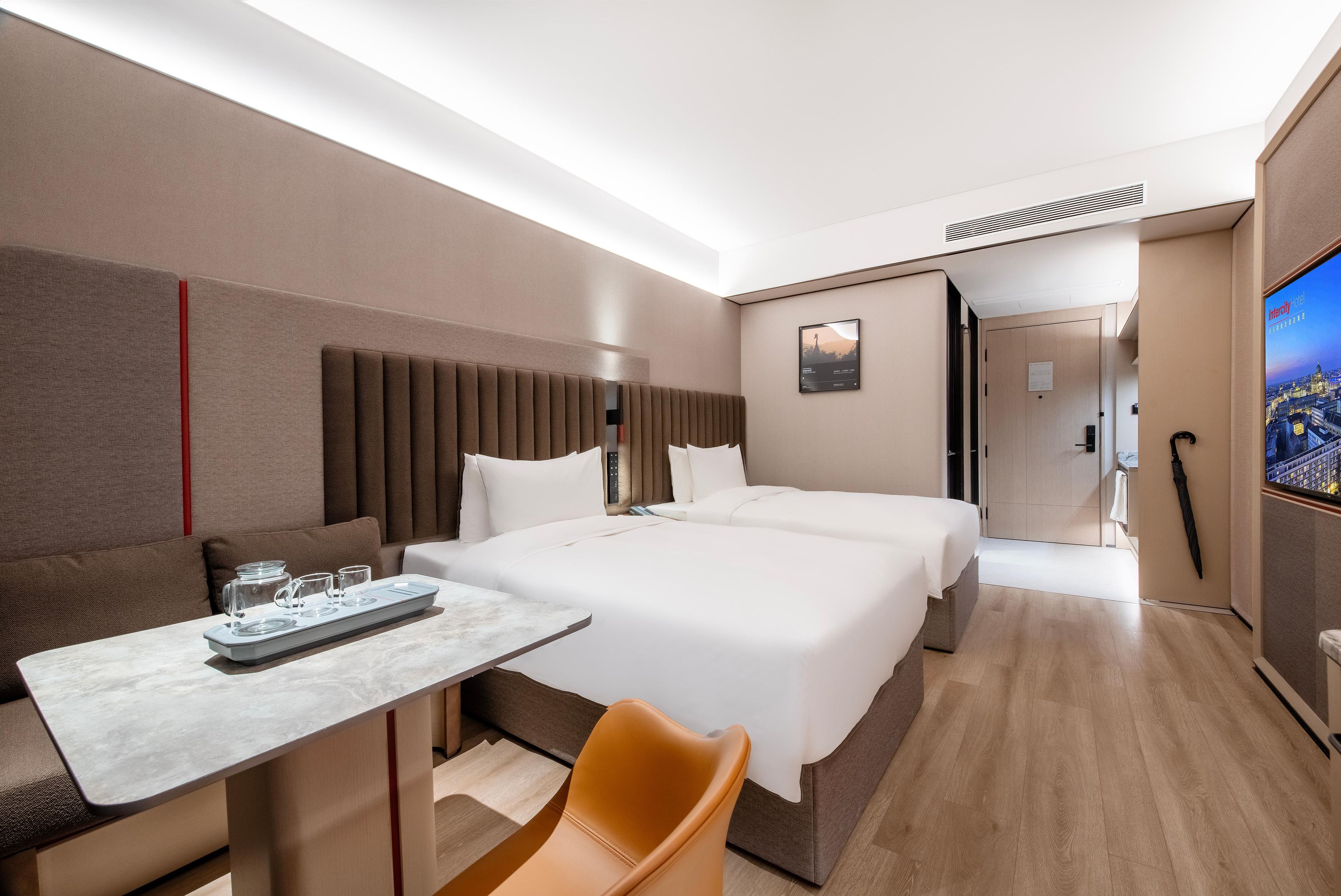 A modern hotel room with a large bed, a sofa, a small table with glasses, and a TV screen displaying a cityscape.