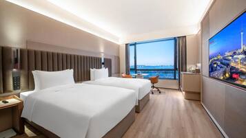 A hotel room with two beds, a large window with a city view, a flat-screen TV, and a small table.